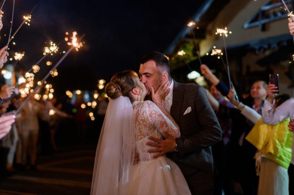 Side view of beautiful bride and bridegroom, which hugging and kissing each other while guests holding cold sparkles which burning and creating glowing arch during night ceremony outdoor